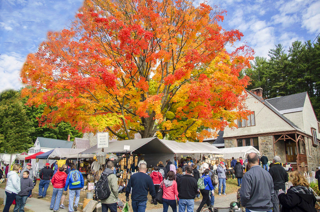 Crowds shopping vendor tents beneath a bright orange maple at the World's Largest Garage Sale in Warrensburg, NY